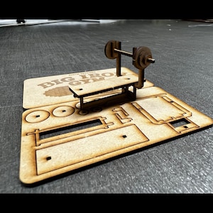 Peut inclure: Un modèle en bois d'un banc de musculation avec une barre et des poids. Le modèle est fait de bois découpé au laser et est conçu pour être assemblé. Le modèle est intitulé "Big Iron Gym".