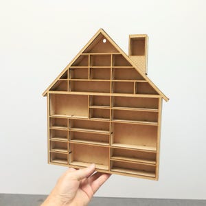 May include: A wooden house-shaped shelf with multiple compartments and a chimney. The shelf is made of light brown wood and has a triangular roof. The design includes various sized rectangular shelves, perfect for displaying small items.