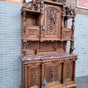 May include: A large, intricately carved wooden cabinet with a marble top. The cabinet has two doors on the bottom and a glass-fronted cabinet on top. The cabinet is decorated with ornate carvings of flowers, leaves, and other designs.