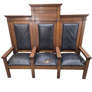 May include: A three-seat wooden bench featuring a high backrest with a central raised panel. The bench has dark leather upholstery on the backrests and seat cushions, which show signs of wear and tear, and includes wooden armrests and legs.