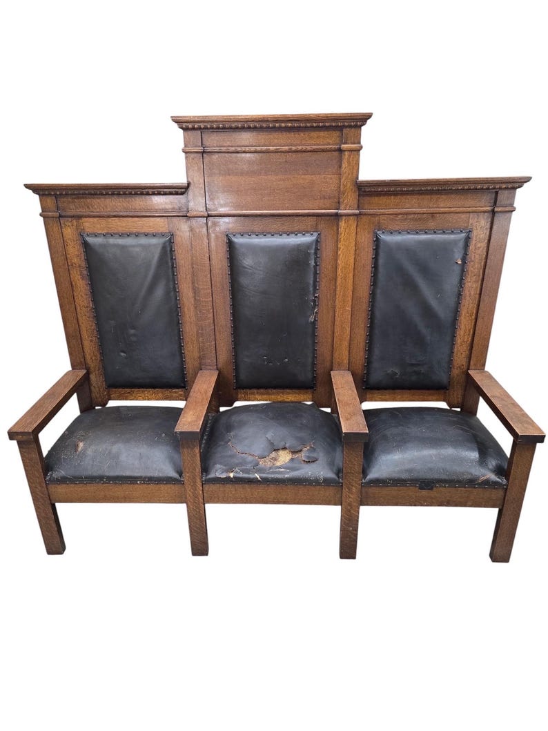 May include: A three-seat wooden bench featuring a high backrest with a central raised panel. The bench has dark leather upholstery on the backrests and seat cushions, which show signs of wear and tear, and includes wooden armrests and legs.
