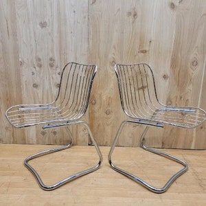 May include: Two chrome wire chairs with a curved, open back and a simple, curved seat. The chairs are on a wooden floor.