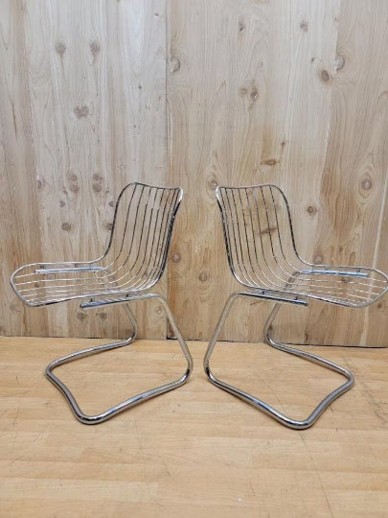 May include: Two chrome wire chairs with a curved, open back and a simple, curved seat. The chairs are on a wooden floor.