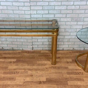 May include: A set of two gold-toned glass tables with a modern design. The larger table is rectangular with a glass top and a gold-toned metal frame. The smaller table is round with a glass top and a gold-toned metal frame.