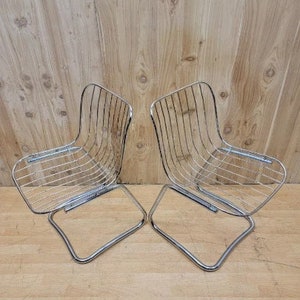May include: Two chrome wire chairs with a curved back and a square seat. The chairs are on a wooden floor.