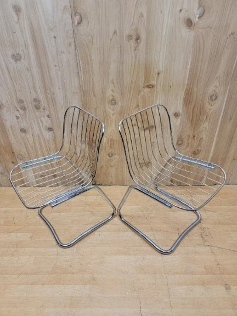 May include: Two chrome wire chairs with a curved back and a square seat. The chairs are on a wooden floor.