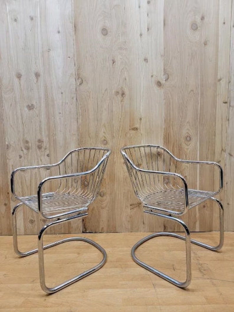 May include: Two chrome wire chairs with a curved back and a curved seat. The chairs are on a wooden floor.
