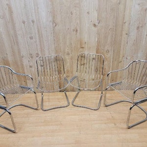 May include: Four chrome wire chairs with a curved back and a curved seat. The chairs are arranged in a row on a wooden floor.