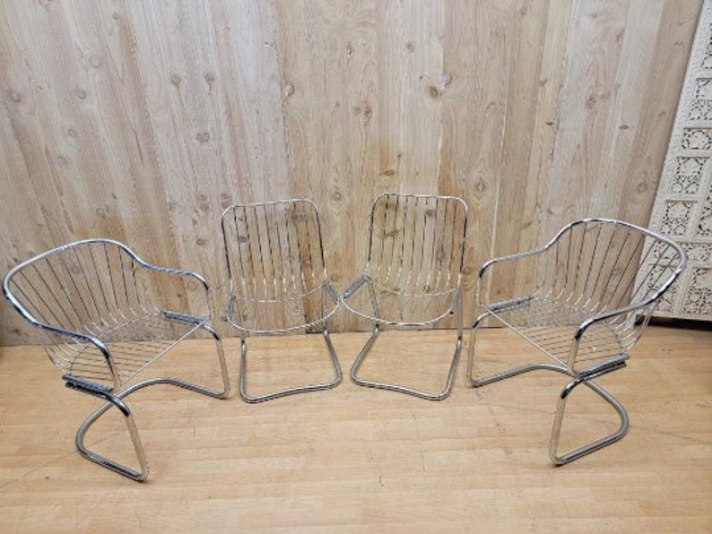 May include: Four chrome wire chairs with a curved back and a curved seat. The chairs are arranged in a row on a wooden floor.