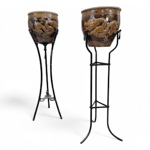 Puede incluir: Dos macetas de cerámica marrón con diseños de dragones sobre soportes de metal negro. Las macetas tienen un acabado texturizado y un borde marrón claro. Los soportes tienen un diseño curvo y delgado.