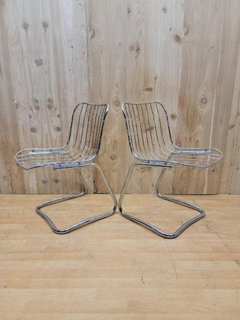May include: Two chrome wire chairs with a curved back and a simple design. The chairs are on a wooden floor.