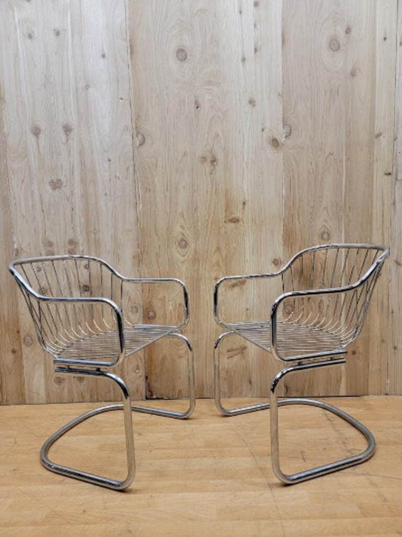May include: Two chrome wire chairs with a curved back and a curved seat. The chairs are on a wood floor.