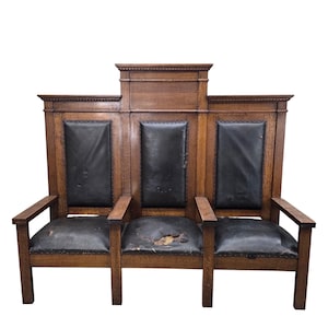 May include: A three-seat wooden bench featuring a high backrest with three distinct sections. The frame is constructed from brown wood with a visible grain. Each seat and back panel is upholstered in black material, showing signs of wear.