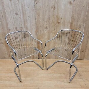 May include: Two chrome wire chairs with a curved back and a wire seat. The chairs are on a wooden floor.