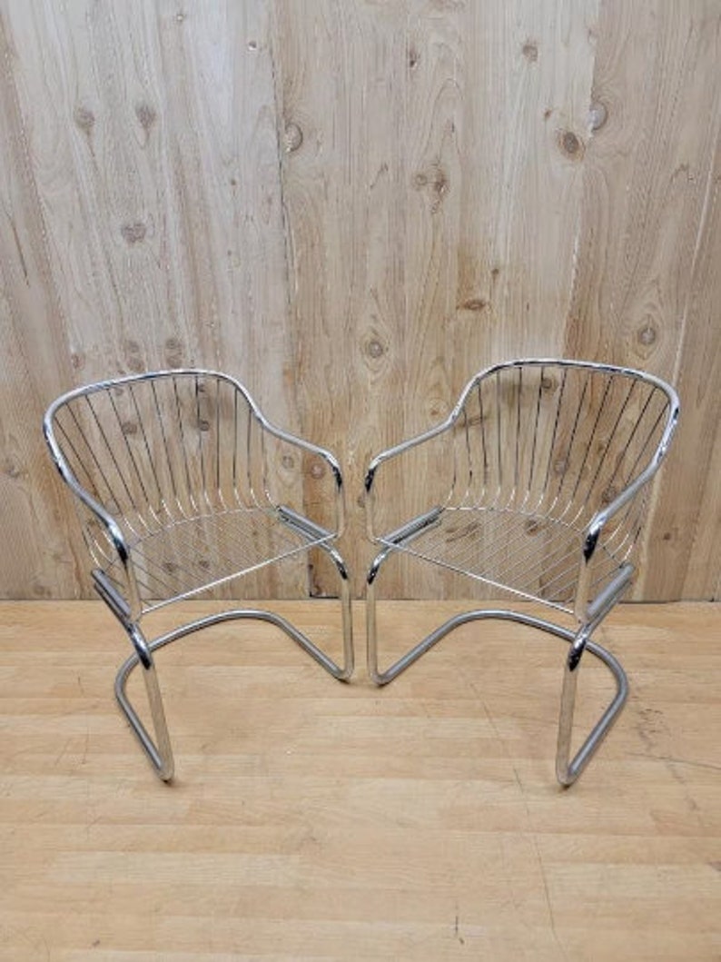 May include: Two chrome wire chairs with a curved back and a wire seat. The chairs are on a wooden floor.