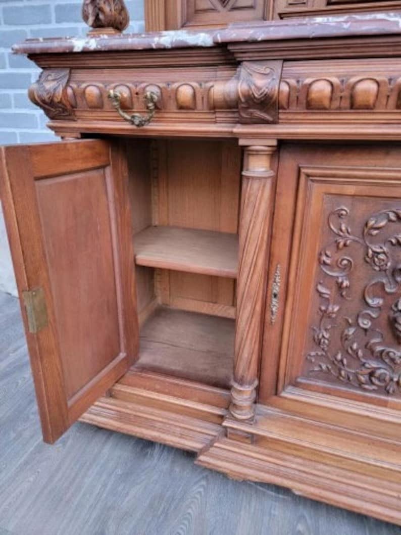 May include: A large, antique wooden cabinet with intricate carvings and a marble top. The cabinet has two doors, one of which is open, revealing shelves inside. The cabinet is made of dark wood and has a rich, warm patina.