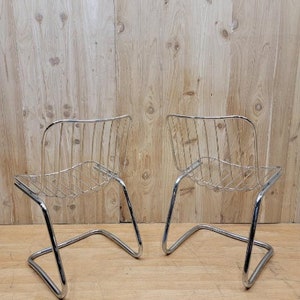 May include: Two chrome wire chairs with a curved back and a simple design. The chairs are on a wooden floor.