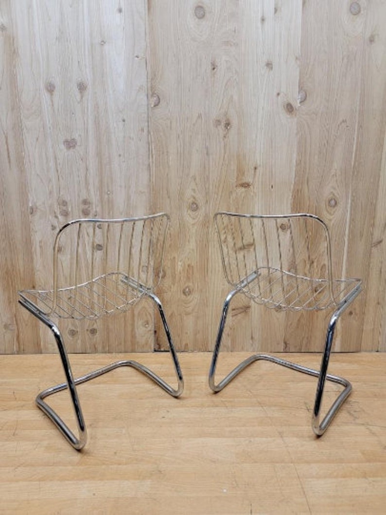 May include: Two chrome wire chairs with a curved back and a simple design. The chairs are on a wooden floor.