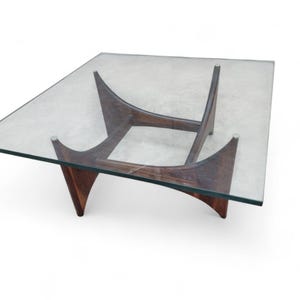 Op de afbeelding: Vierkante salontafel met een helder glazen blad en een donkerhouten onderstel. Het onderstel heeft een uniek ontwerp met gebogen steunen en een mid-century modern esthetiek. De tafel is ongeveer 91 cm vierkant.