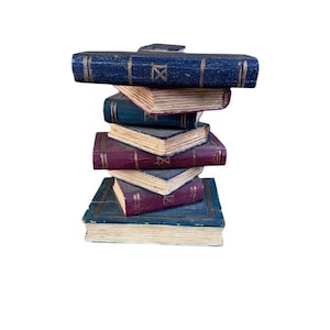 Puede incluir: Una pila de seis libros decorativos con cubiertas azules y burdeos. Los libros están dispuestos en una torre precaria, con los lomos y los bordes visibles. Cada libro tiene un diseño dorado en el lomo.