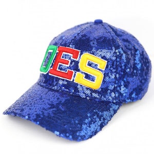 May include: A blue sequined baseball cap with the word "DOES" embroidered on the front in green, red, and yellow. The cap has a curved brim and a button on top. The sequins give the cap a shiny appearance.