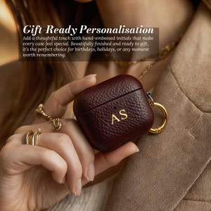 May include: A burgundy leather AirPod case with gold-colored hardware and the initials "AS" embossed on the front. The case has a textured surface and is held by a person. The text "Gift-Ready Personalisation" is visible in the background.