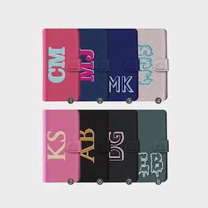 Pode incluir: Oito capas de telefone diferentes com iniciais personalizadas. As capas são de várias cores, incluindo rosa, preto, azul e verde. As iniciais são bordadas em diferentes cores e estilos.