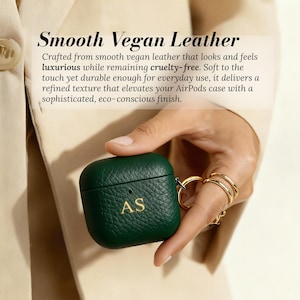 May include: A dark green AirPods case made of smooth vegan leather. The case has a textured surface and gold-colored initials "AS". The case is held by a person wearing gold rings. The text "Smooth Vegan Leather" is at the top.