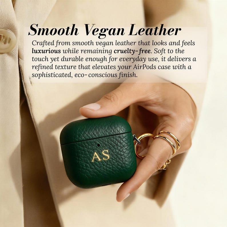 May include: A dark green AirPods case made of smooth vegan leather. The case has a textured surface and gold-colored initials "AS". The case is held by a person wearing gold rings. The text "Smooth Vegan Leather" is at the top.