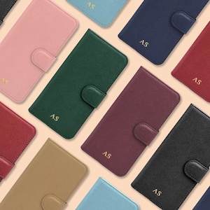 Puede incluir: Una colección de fundas de teléfono coloridas en varios tonos, incluyendo rosa, verde, azul marino, rojo y negro. Cada funda presenta un cierre de botón a presión y las iniciales "AS" en letras doradas. Las fundas están dispuestas sobre un fondo beige claro.