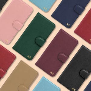 May include: A selection of phone cases in a variety of colours, such as pink, green, and blue. Each case has a snap closure and the initials "AS" in gold. The cases are displayed on a light beige background.