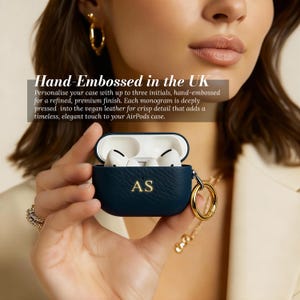 May include: A navy blue AirPods case with the initials "AS" in gold lettering. The case is open, revealing white earbuds. The text "Hand-Embossed in the UK" is visible. A gold-colored ring and chain are attached to the case.