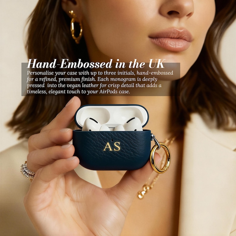 May include: A navy blue AirPods case with the initials "AS" in gold lettering. The case is open, revealing white earbuds. The text "Hand-Embossed in the UK" is visible. A gold-colored ring and chain are attached to the case.