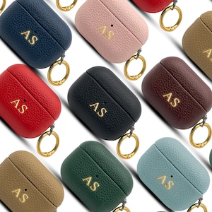 May include: A collection of personalized AirPod cases in various colors, including navy, pink, red, black, brown, green, and teal. Each case features the gold-colored initials "AS" and a gold-tone keyring.