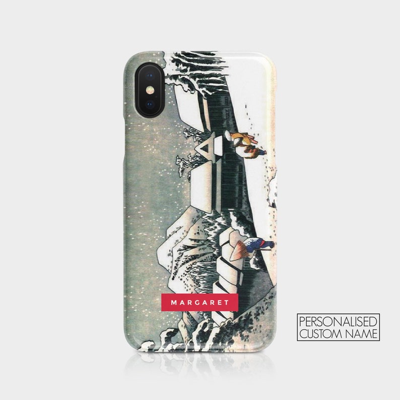 May include: A white iPhone case with a Japanese-style illustration of a snowy mountain scene with two figures walking on a path. The text "MARGARET" is printed in red at the bottom of the case. The text "PERSONALISED CUSTOM NAME" is printed in white on a black background in the lower right corner.