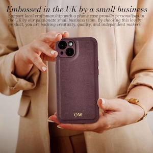 May include: A burgundy leather phone case with a gold embossed monogram "OW". The case is designed for an iPhone with a camera bump.