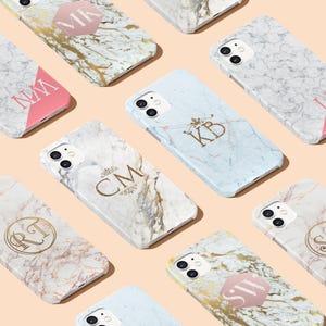 Op de afbeelding: Assortiment van marmeren telefoonhoesjes met gouden en roze accenten. Elk hoesje heeft een ander monogram met een decoratief ontwerp. De hoesjes worden gepresenteerd op een lichtroze achtergrond.