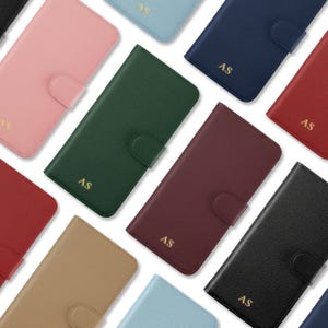 Könnte beinhalten: Eine Sammlung von Leder-Handyhüllen in verschiedenen Farben, darunter Rosa, Grün, Marineblau und Rot. Jede Hülle hat einen Schnappverschluss und die Initialen "AS" in goldfarbenen Buchstaben. Die Hüllen sind auf weißem Hintergrund angeordnet.