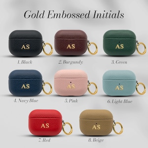 May include: A collection of eight leather AirPod cases in various colors, including black, burgundy, green, navy blue, pink, light blue, red, and beige. Each case features gold embossed initials and a gold-colored ring.