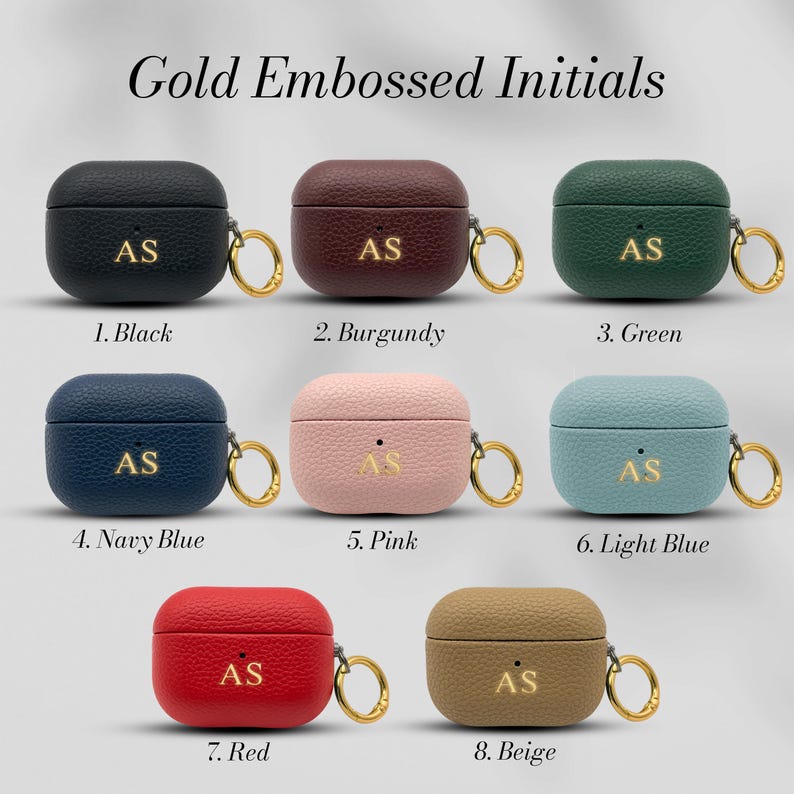 May include: A collection of eight leather AirPod cases in various colors, including black, burgundy, green, navy blue, pink, light blue, red, and beige. Each case features gold embossed initials and a gold-colored ring.