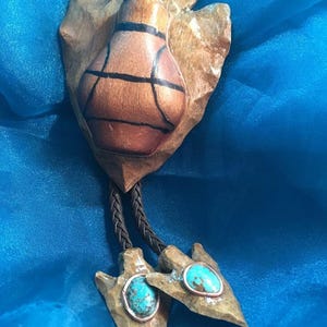 May include: A bolo tie featuring a brown, copper-toned pendant with black lines and a turquoise stone. The pendant is attached to a brown leather cord with two arrowhead-shaped accents, each with a turquoise stone and copper trim.