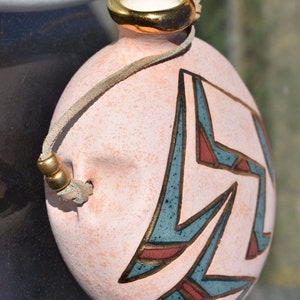 Pode incluir: Uma garrafa de cerâmica rosa com borda dourada e alça de couro. A garrafa apresenta um design geométrico em azul-petróleo, vermelho e dourado. Um item decorativo único.