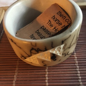 Puede incluir: Un pequeño cuenco de cerámica blanquecina con una superficie texturizada y marcas oscuras. Dentro del cuenco hay un trozo de papel doblado con texto impreso. El cuenco está sobre una esterilla de bambú marrón.