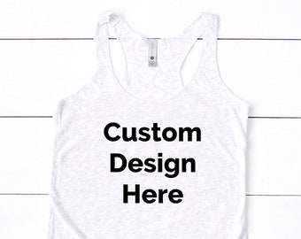 May include: A white racerback tank top with the text "Custom Design Here" in bold black letters. The tank top is made of a soft, light-colored fabric and is laying on a white wooden surface. A small circular logo is in the bottom right corner.