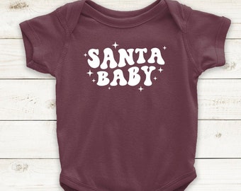 May include: A maroon baby bodysuit featuring the white text "SANTA BABY" in a retro font, with small white starbursts. The short-sleeved onesie has poppers at the bottom. A festive garment for a baby.