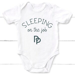 May include: White baby onesie with short sleeves and the text "SLEEPING on the job" in black. A stylized black and white "DP" logo is below the text.