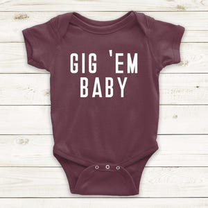 May include: A maroon baby bodysuit with the white text "GIG 'EM BABY". The bodysuit has short sleeves and three poppers at the bottom. The background is a light wood grain pattern.