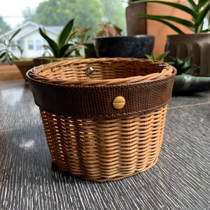 May include: A small, round wicker basket with a brown leather trim and a gold metal button. The basket is sitting on a dark wood surface.
