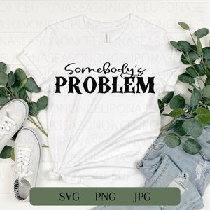 以下が含まれることがあります： 白いTシャツに黒い文字で「Somebody's PROBLEM」と書かれています。Tシャツは折り畳まれて、白い背景に緑の植物と白いスニーカーと一緒に置かれています。