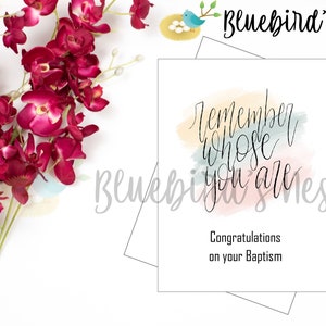 May include: A white greeting card with the text "Remember whose you are" in elegant script, with watercolor accents. The card is for a baptism, with the words "Congratulations on your Baptism" below. Burgundy flowers are in the background.